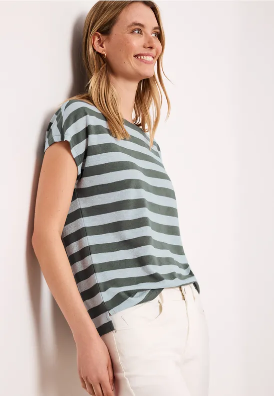 Een vrouw leunt tegen een muur, draagt een groen-grijs gestreept T-shirt met korte mouwen en een lichte broek.