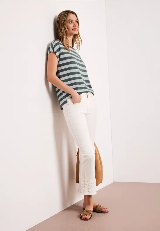 Vrouw leunt tegen muur, draagt groen-wit gestreept shirt, witte flared broek met franjes en sandalen.