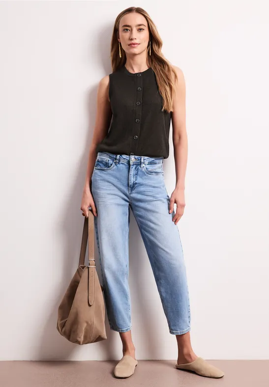 Vrouw draagt zwarte mouwloze top, lichtblauwe jeans, beige instappers en grote tas tegen witte achtergrond.