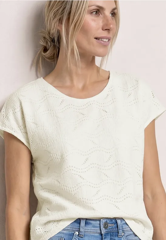 Vrouw met een wit, kortgebreid shirt met golvend patroon, gecombineerd met een spijkerbroek.