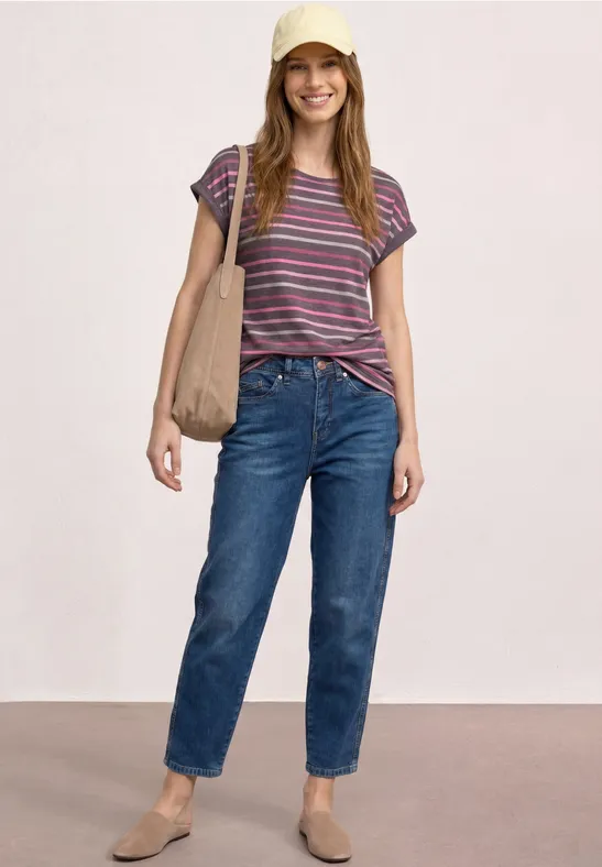 Vrouw staat voor lichte achtergrond, draagt jeans, gestreept shirt, platte schoenen en schoudertas.