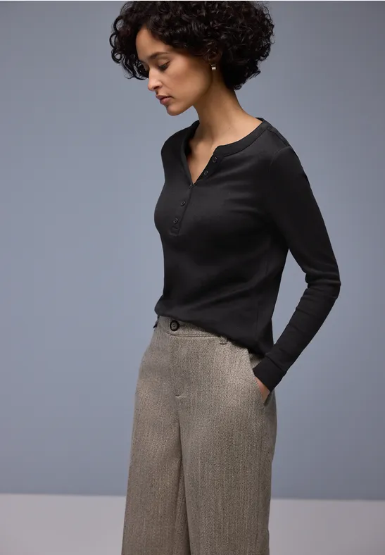 Person in schwarzem Langarmshirt mit V-Ausschnitt und grauen, taillierten Hosen vor neutralem Hintergrund.