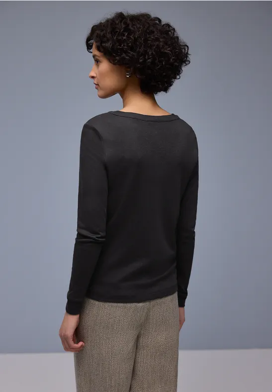 Person von hinten in schwarzem Langarmshirt und beigen Hosen, lockige Haare, neutraler Hintergrund.