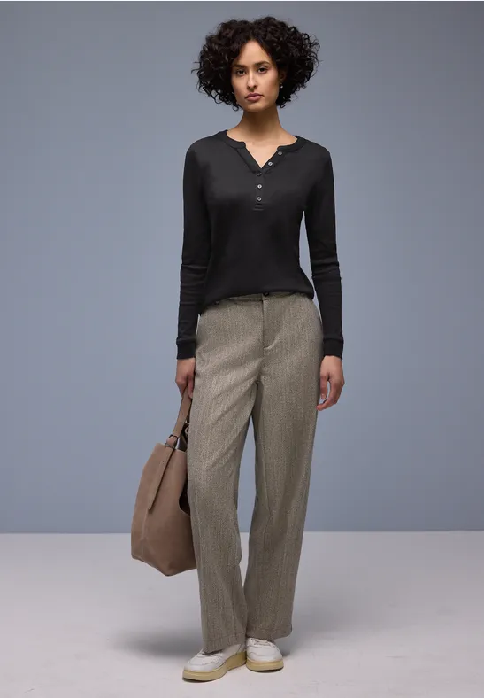 Frau in schwarzem Langarmtop, weiten grauen Hosen, weißen Schuhen und großer Handtasche.