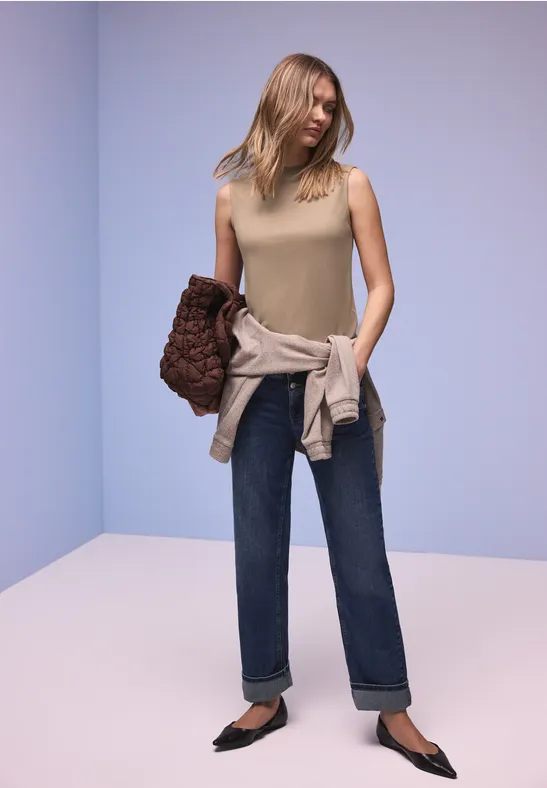Frau trägt beiges Top, Jeans, schwarze spitze Schuhe und hält eine braune Clutch vor hellem Hintergrund.