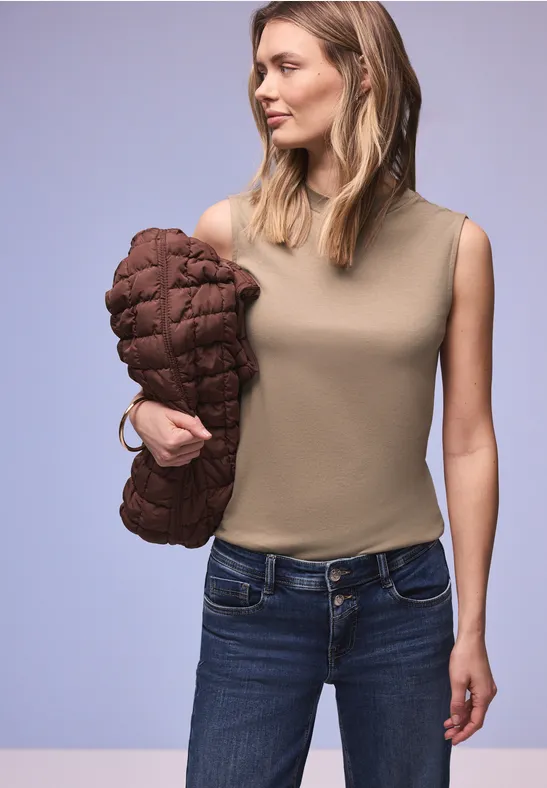 Eine Person in ärmellosem Oberteil und Jeans hält eine braune Steppjacke vor schlichtem Hintergrund.