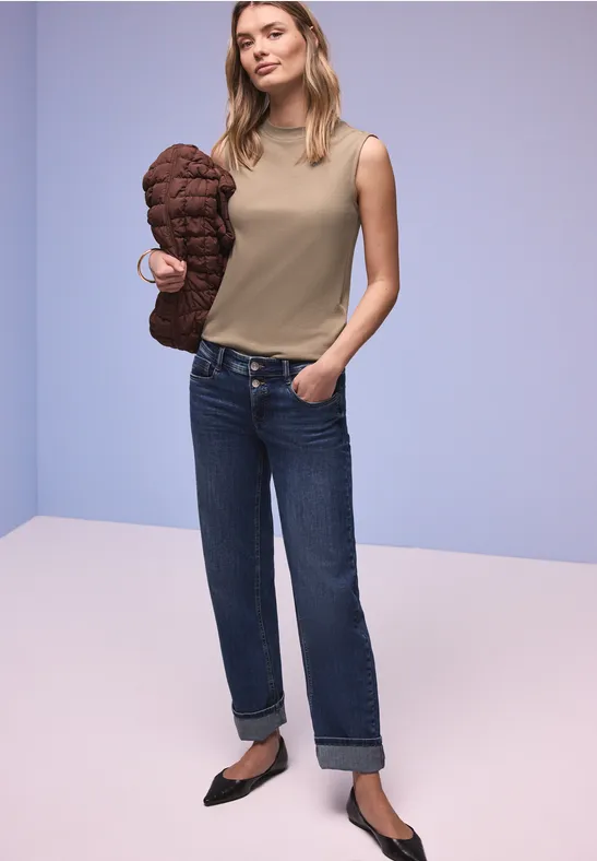 Frau in ärmellosem Top, geraden Jeans und Ballerinas hält eine gesteppte Jacke vor schlichtem Hintergrund.