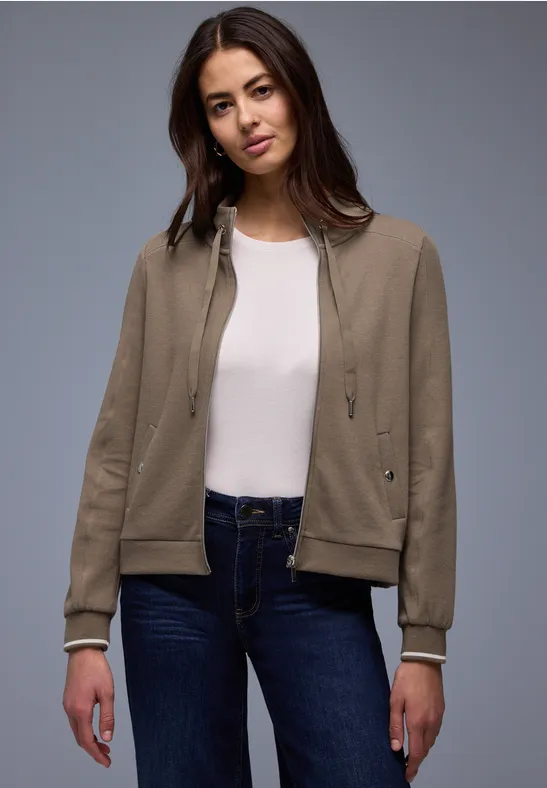 Frau in beiger Jacke, weißem Shirt und dunklen Jeans vor grauem Hintergrund.