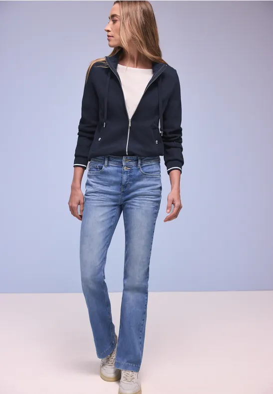 Person in Kapuzenjacke, weißem Shirt, blauen Jeans und weißen Sneakern vor neutralem Hintergrund.