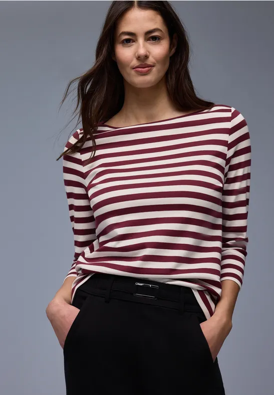 Frau in gestreiftem Langarmshirt und dunklen Hosen steht mit Händen in den Taschen vor grauem Hintergrund.