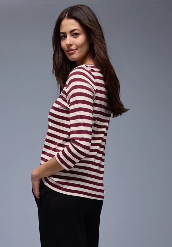 Frau im Profil mit langem braunem Haar trägt ein weiß-rot gestreiftes 3/4-Arm-Shirt und schwarze Hose.