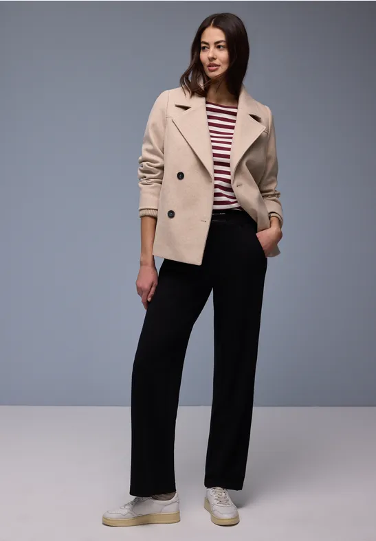 Eine Person in einer beigen Jacke, gestreiftem Shirt und schwarzen Weithosen vor grauem Hintergrund.