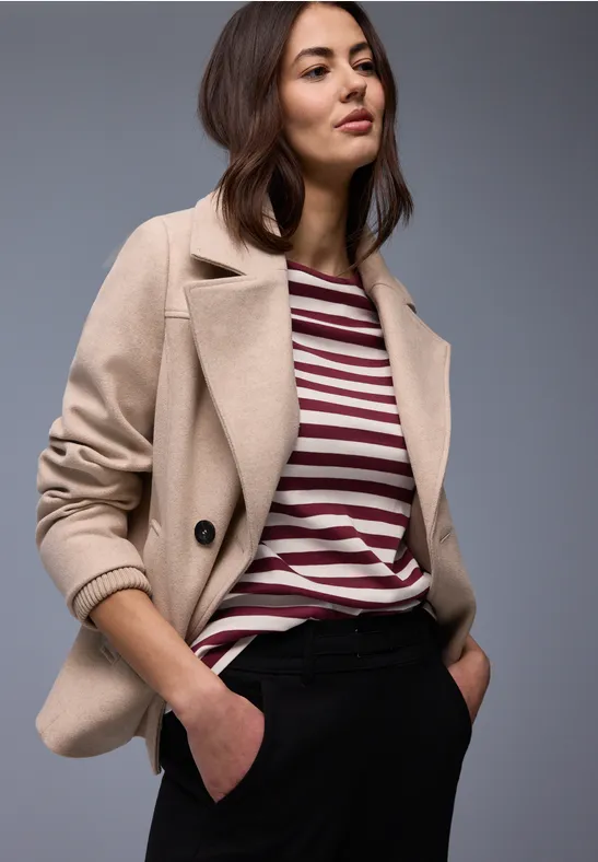 Frau trägt beige Blazer, gestreiftes Shirt und dunkle Hose vor neutralem Hintergrund.