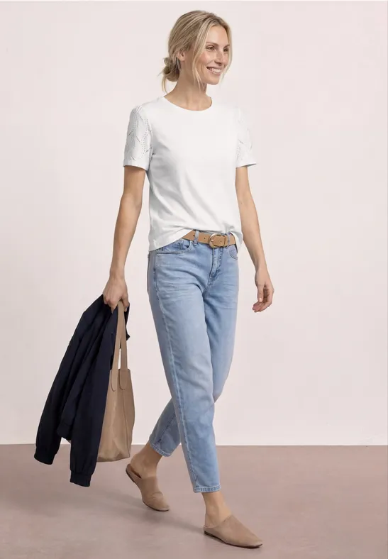 Frau trägt weißes T-Shirt, blaue Jeans und beigen Mules, hält eine dunkelblaue Jacke und Tasche.
