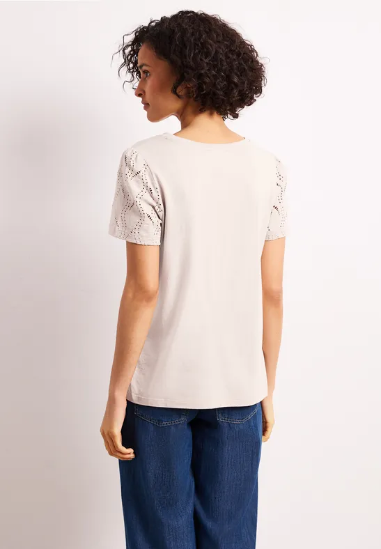 Vrouw van achteren in een licht beige t-shirt met kanten mouwen en een blauwe spijkerbroek tegen een rustige achtergrond.