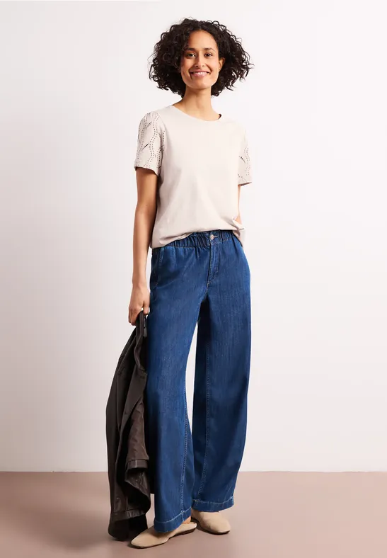 Vrouw met beige t-shirt en wijde blauwe jeans, houdt een zwart jack vast, lichte schoenen, neutrale achtergrond.