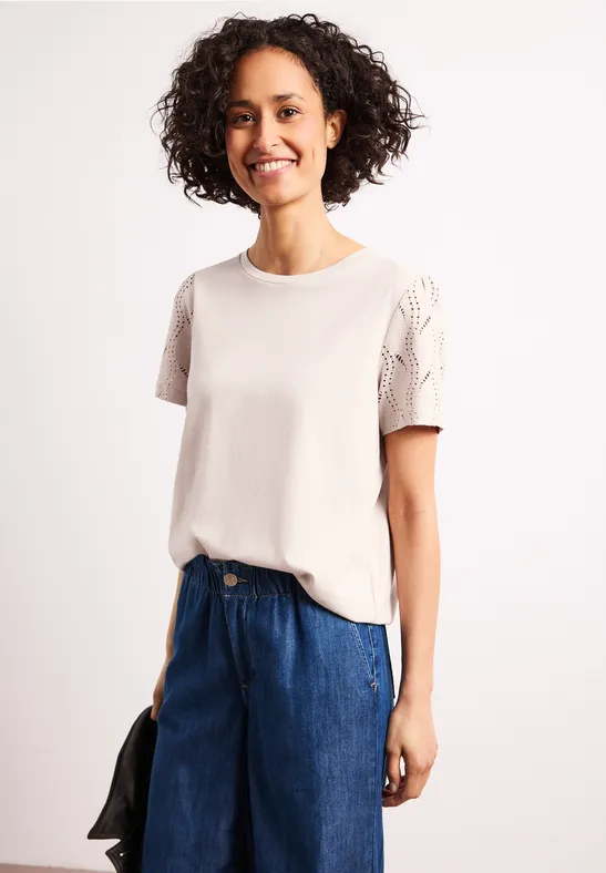 Vrouw in wit t-shirt met kanten mouwen en blauwe jeans, houdt een zwarte jas in haar hand.