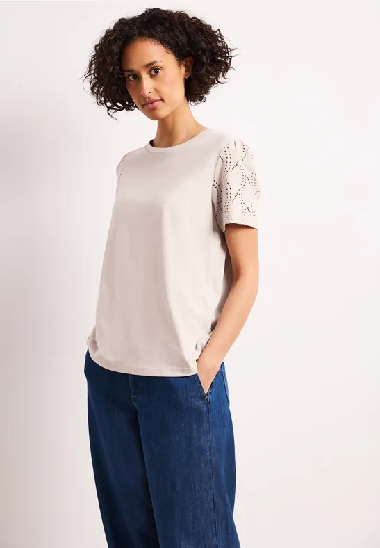 Vrouw in crèmekleurig T-shirt met kantmouwen en wijde blauwe jeans, handen in de zakken.