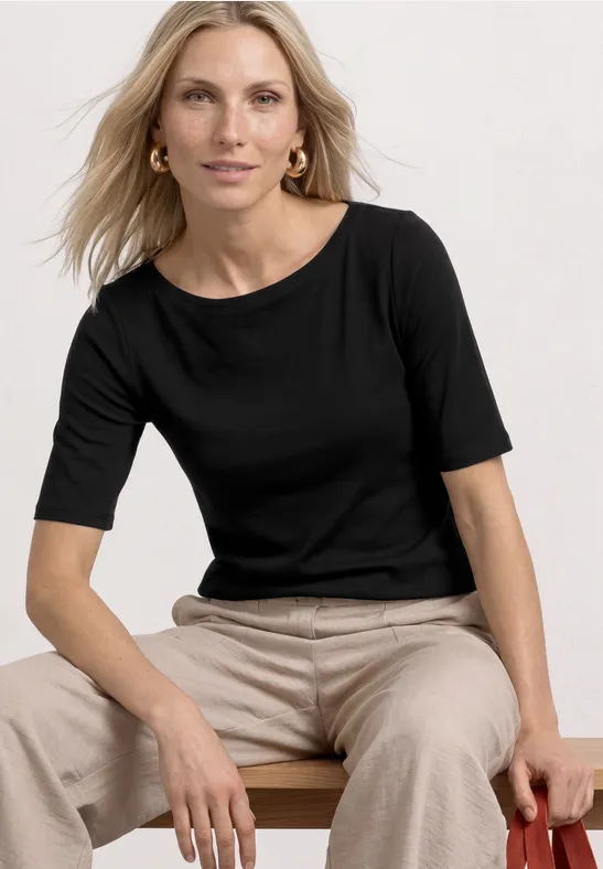Une femme en haut noir à manches mi-longues et pantalon beige est assise sur un tabouret devant un fond clair.