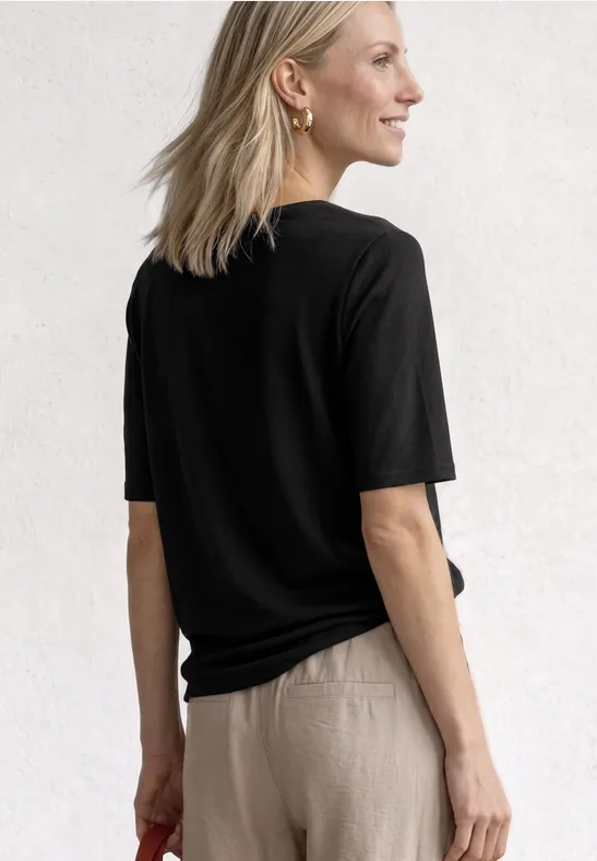 Femme vue de dos, portant un t-shirt noir et un pantalon beige sur fond neutre.