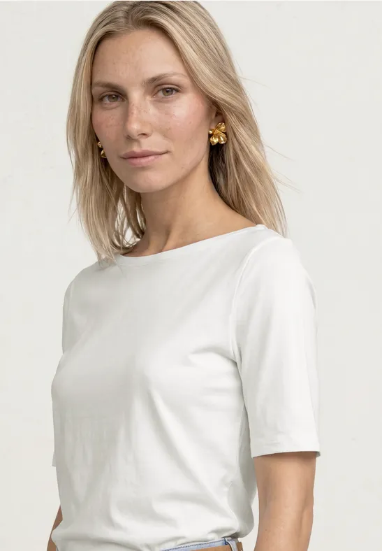 Femme portant un t-shirt blanc ample et des boucles d'oreilles dorées sur fond clair.