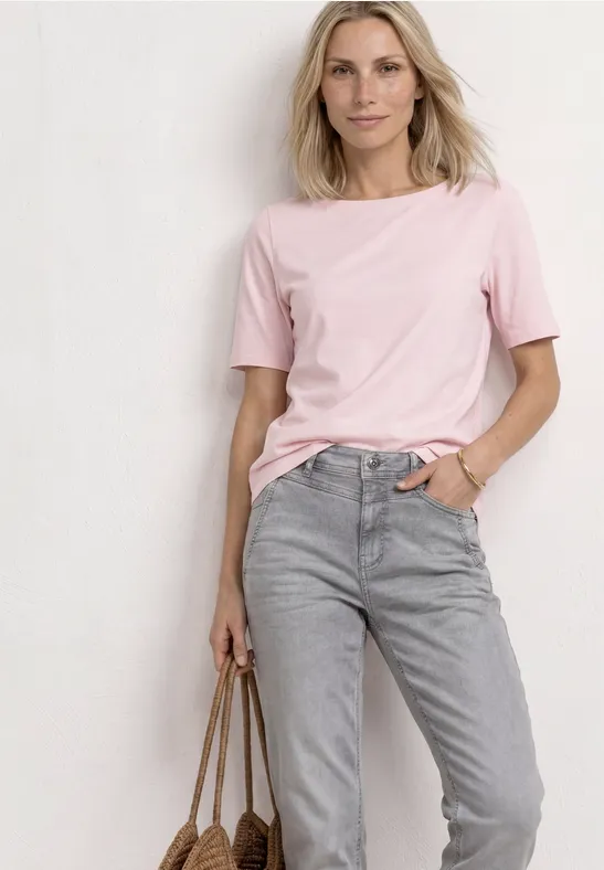 Frau in rosa T-Shirt und grauen Jeans hält eine Tasche in der Hand, vor weißem Hintergrund.