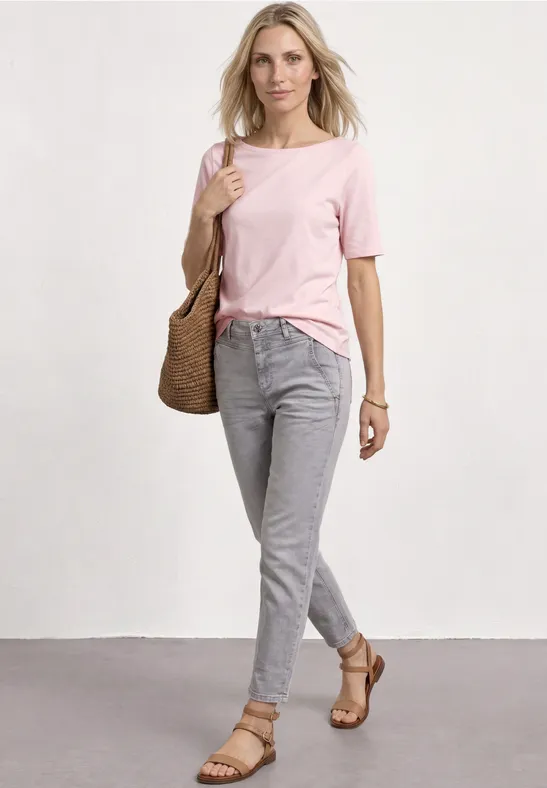 Frau in rosa T-Shirt, grauen Hosen, geflochtener Tasche und Sandalen, vor weißer Wand.