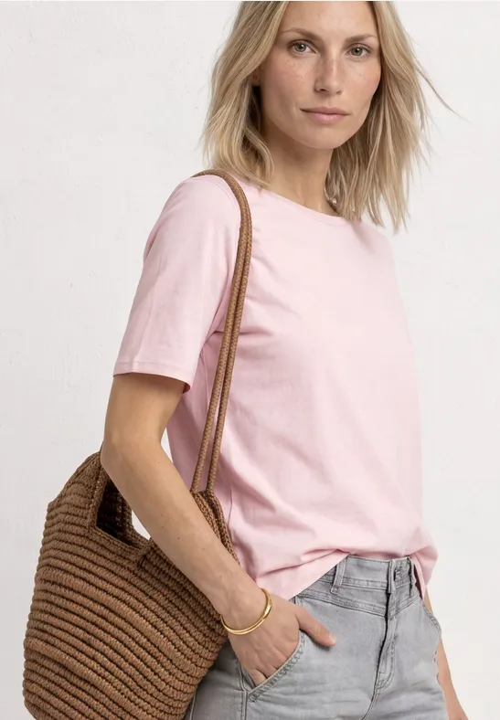 Frau trägt rosa T-Shirt, graue Hose und eine geflochtene Strohtasche über der Schulter.