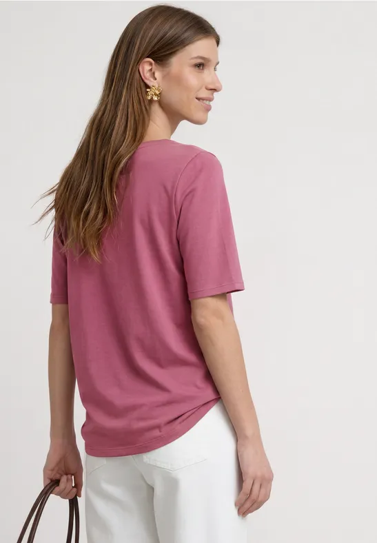 Frau von hinten in rosafarbenem T-Shirt, weißer Hose, braunen Haaren und goldener Kette.