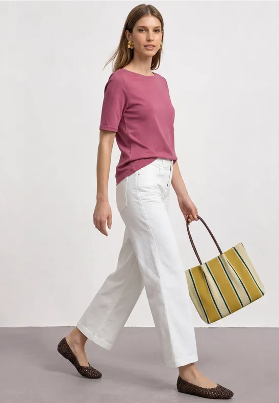 Frau in rosa T-Shirt, weißer Hose und braunen Loafers trägt gestreifte Tote-Bag vor neutralem Hintergrund.
