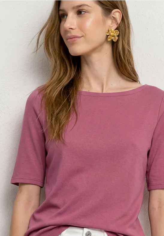 Frau mit rosa Kurzarmshirt, großer goldener Ohrring, Gesicht unscharf, vor heller Wand.