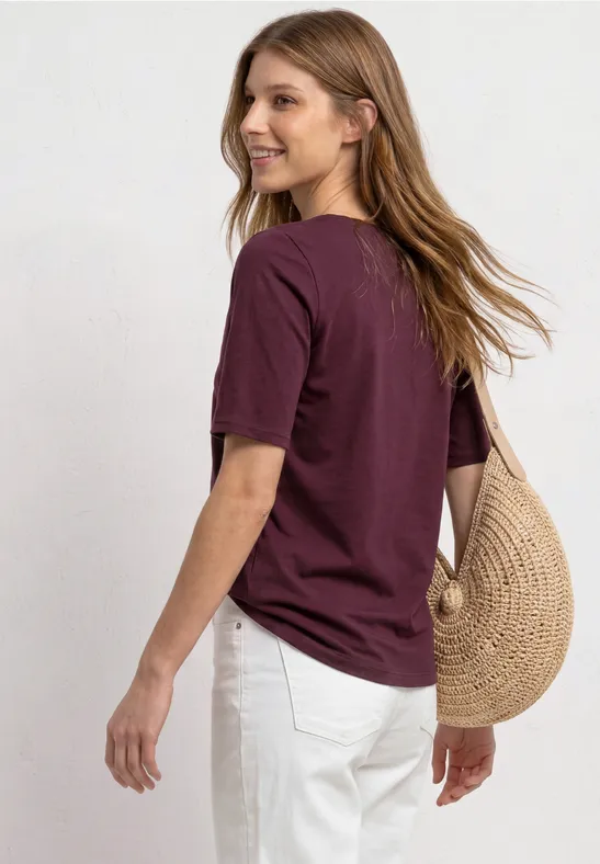 Vrouw in bordeaux T-shirt en witte broek draagt een ronde strooien tas over haar schouder.