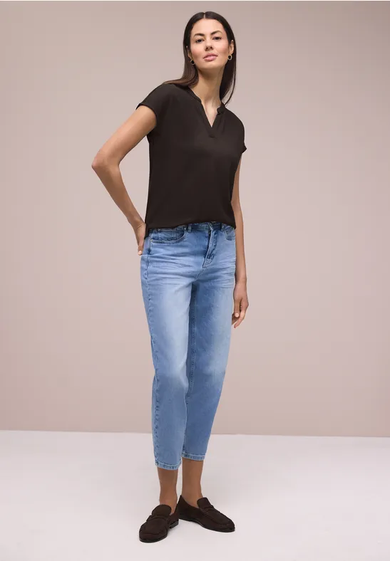 Frau in schwarzem T-Shirt, blauen Jeans und schwarzen Loafers vor schlichter Kulisse stehend.