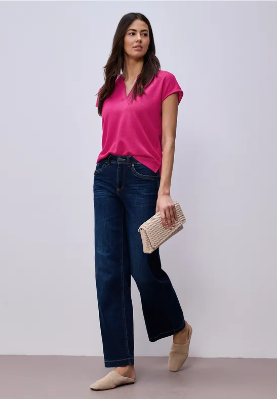 Frau in pinker Bluse, weiten Jeans und beigen Ballerinas, hält eine beige Clutch.