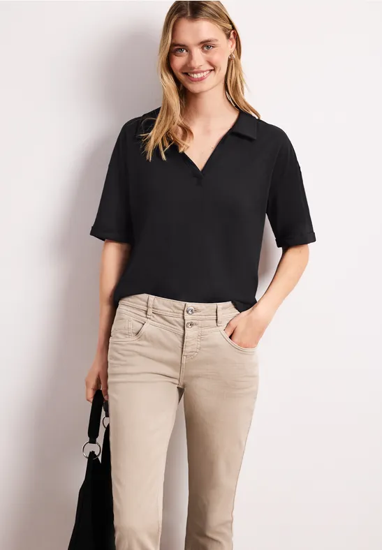 Eine Frau trägt eine schwarze V-Ausschnitt-Bluse und beigefarbene Hose, hält eine kleine schwarze Handtasche.