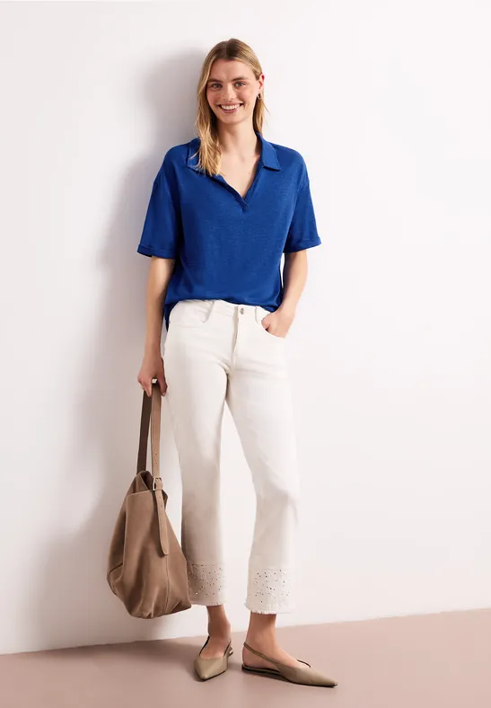 Vrouw in blauw shirt, witte broek en beige tas voor een lichte achtergrond