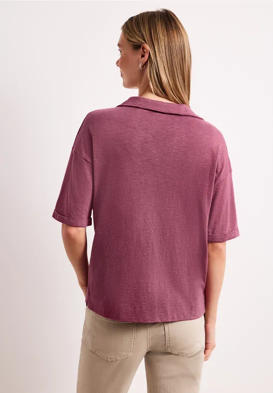 Vrouw van achteren in een lichtpaarse korte blouse en beige broek tegen witte achtergrond.