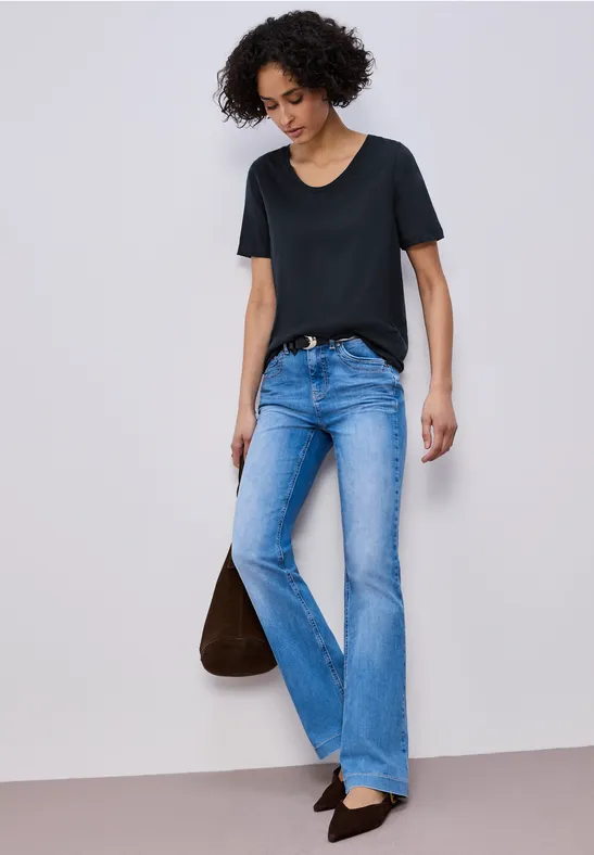Frau in schwarzem T-Shirt, hellblauen Schlagjeans und schwarzen Schuhen hält eine braune Tasche.