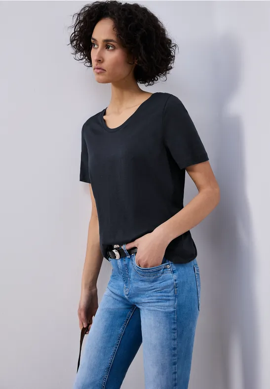 Frau in schwarzem T-Shirt und blauen Jeans seitlich stehend, Hand in der Tasche.