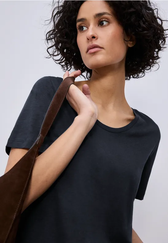 Person mit lockigem Haar trägt schwarzes T-Shirt und hält braune Tasche über der Schulter.