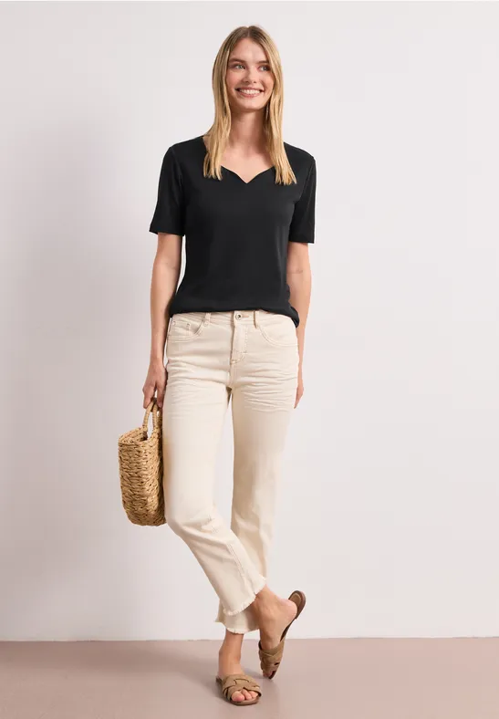 Vrouw met zwart V-hals T-shirt, lichte jeans, bruine loafers en een mandtas voor een neutrale achtergrond.