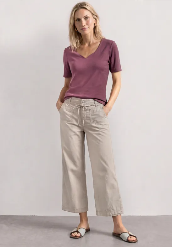 Femme en t-shirt violet et pantalon ample clair devant un fond neutre.