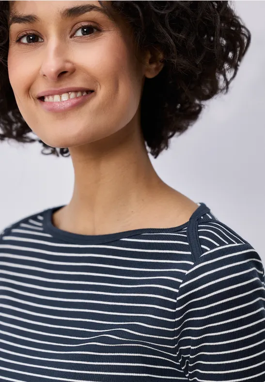 Oberkörper mit kurzen lockigen dunklen Haaren, trägt ein blau-weiß gestreiftes Shirt vor hellem Hintergrund.