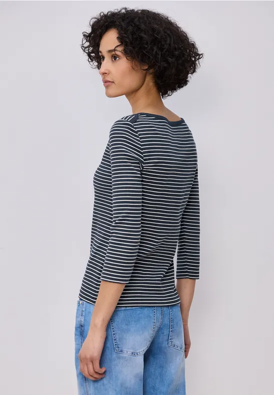Person mit lockigem Haar in blau-weiß gestreiftem Oberteil und hellblauen Jeans, seitlich stehend.