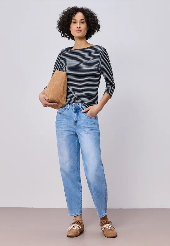 Eine Person in grauem Langarmshirt, hellblauen Jeans und braunen Halbschuhen hält eine Papiertüte in der Hand.