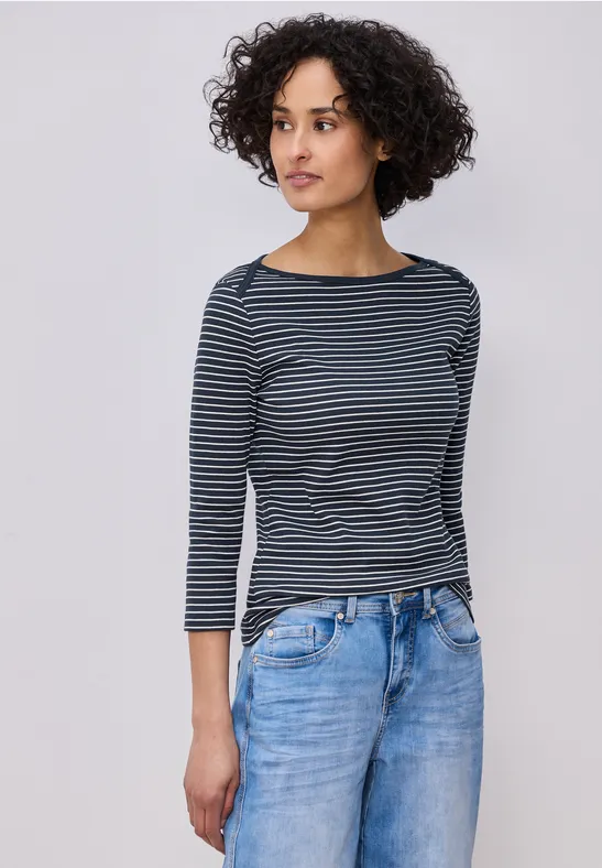 Person mit gestreiftem Langarmshirt und Jeans vor neutralem Hintergrund.