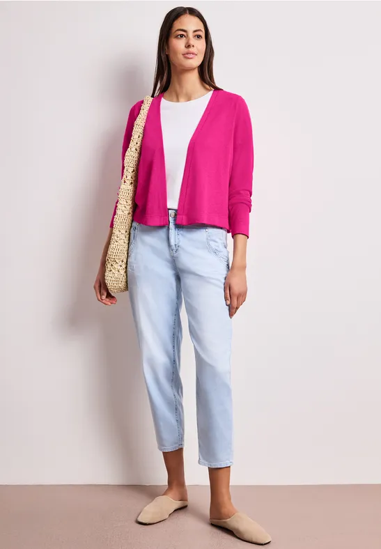 Vrouw in roze vest, witte top, lichtblauwe jeans, beige mules en strooien tas.