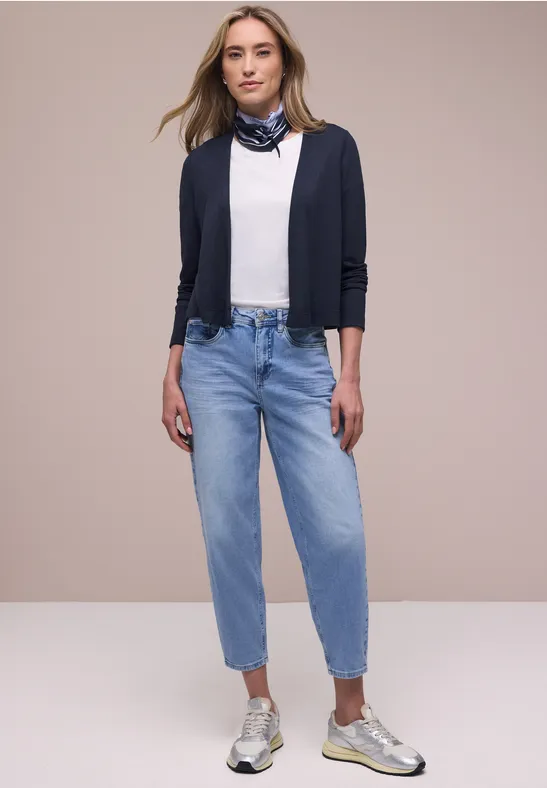 Vrouw met een wit topje, marineblauw vest, lichtblauwe jeans, zilverkleurige sneakers en een sjaaltje.