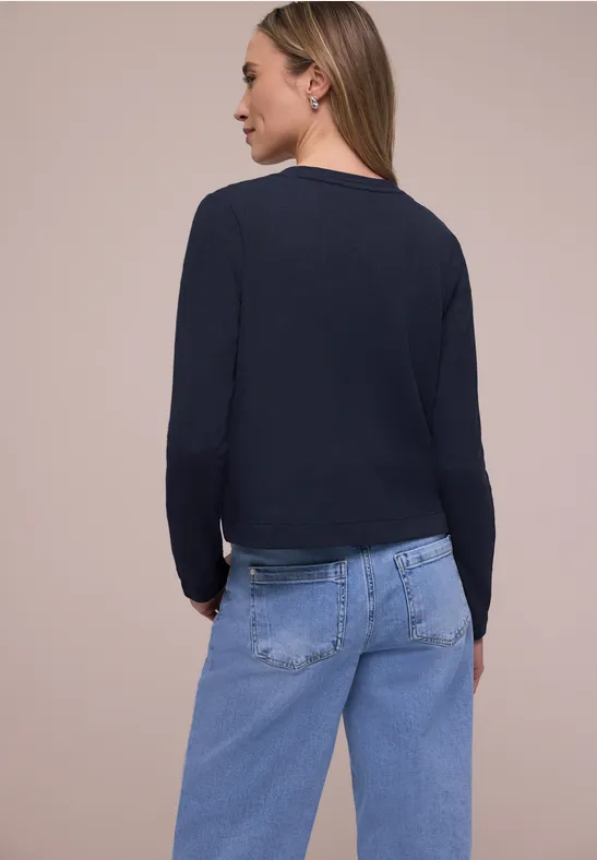 Vrouw van achteren met zwarte top met lange mouwen en blauwe spijkerbroek tegen een neutrale achtergrond.