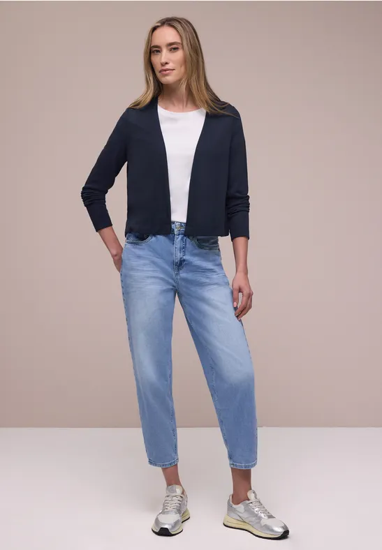 Vrouw in wit t-shirt, donkerblauwe cardigan, lichtblauwe jeans en witte sneakers tegen neutrale achtergrond.